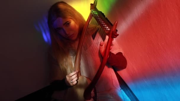 Сверху блондинка играет традиционную музыку на лире, сидя в комнате с разноцветной подсветкой - Кадры, видео