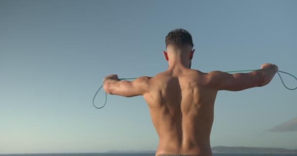 Back view of muscular male bodybuilder with naked torso standing on seashore and doing exercises with resistance band during workout in summer - Footage, Video