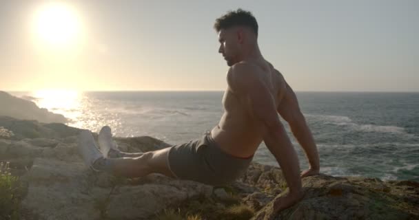 Vista lateral del entrenamiento de atleta masculino sin camisa enfocado en la orilla del mar y haciendo ejercicios de tríceps mientras mira hacia otro lado - Metraje, vídeo