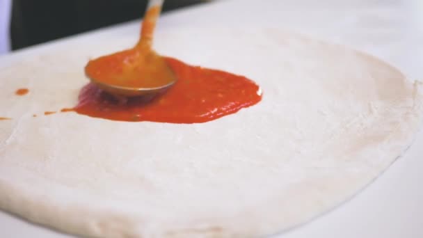 Faceless chef spreading aromatic tomato sauce over dough with big metal spoon while cooking pizza in kitchen - Πλάνα, βίντεο