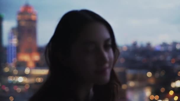 Rauhallinen nainen seisoo lähellä ikkunaa tasainen ja iloisesti katsot kameran hämärtynyt tausta kaupunkikuva illalla - Materiaali, video
