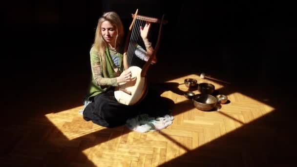 Felülről felnőtt szőke nő ül a napfényben közel éneklő tálak és játszani a hagyományos dallam lant sötét szobában otthon - Felvétel, videó