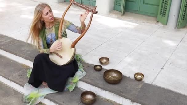 Blonde woman sitting on building stairs near singing bowls and playing lyre on street - Footage, Video
