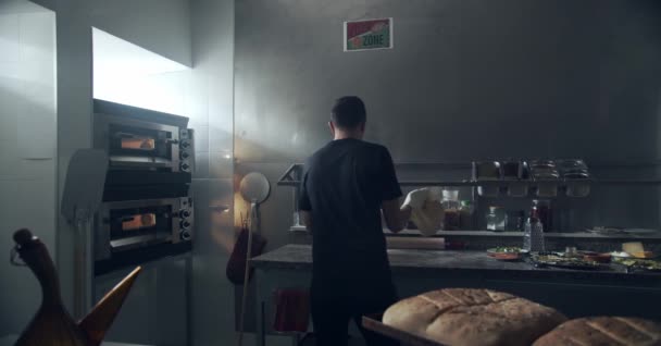 Zadní pohled na člověka převracet a házet protahování čerstvé těsto na mramorovém pultu stolu při přípravě pizzy v blízkosti různých ingrediencí a nádobí v kuchyni pizzerie - Záběry, video