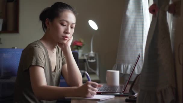 Очаровательная фрилансистка в повседневной одежде, сидящая за столом с ноутбуком и пишущая в блокноте, работая над бизнес-стратегией в домашнем офисе - Кадры, видео