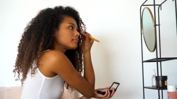 Πλαϊνή άποψη του νεαρού σγουρά μαλλιά εθνοτική θηλυκό εφαρμογή ρουζ από παλέτα στο μάγουλο με καλλυντική βούρτσα μπροστά από έναν καθρέφτη κατά τη διάρκεια της καθημερινής διαδικασίας μακιγιάζ στο σπίτι - Πλάνα, βίντεο
