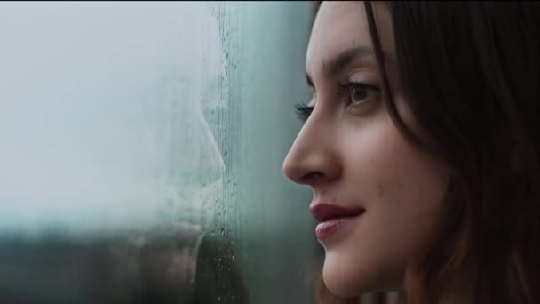 Zijaanzicht van een rustige vrouw die bij het raam staat en thuis door glas regen observeert - Video