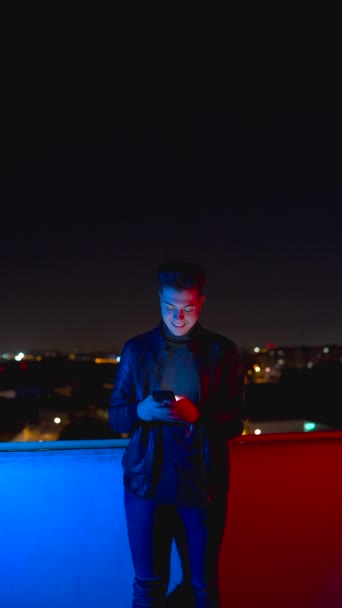 Alegre hombre moderno en chaqueta negra hablando en el teléfono inteligente mientras está de pie en la luz roja y azul en la calle con el cielo azul oscuro en el fondo - Metraje, vídeo