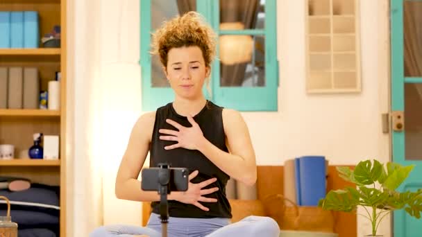 Pozitivní fit žena sedí v lotosu pózovat s rukama na prsou a břiše a provádí hluboké dechové cvičení při nahrávání videa na smartphone - Záběry, video