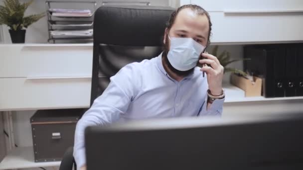 Серьезный ориентированный молодой бизнесмен в медицинской маске обсуждает бизнес-вопрос на мибильном телефоне во время работы с компьютером в современном коворкинг-офисе - Кадры, видео
