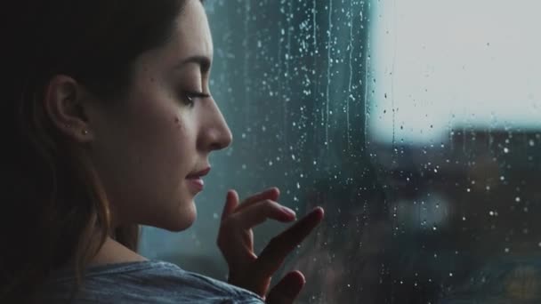 Πλευρική άποψη του ήσυχο θηλυκό στέκεται κοντά στο παράθυρο και παρατηρώντας βροχή μέσα από γυαλί στο σπίτι - Πλάνα, βίντεο