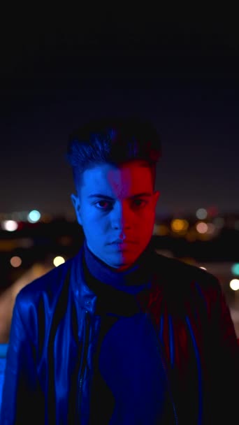 Zeitgenössischer junger Mann, der das halbe Gesicht mit der Hand bedeckt und in die Kamera schaut, während er nachts unter grellem roten und blauen Licht auf verschwommenem Hintergrund der Stadtstraße steht - Filmmaterial, Video