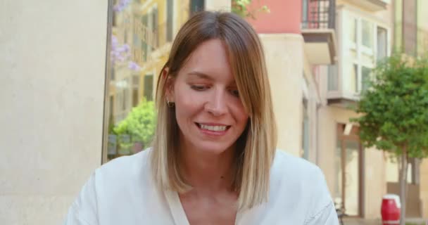 Взрослая женщина в белой рубашке с сочным бургером на обед, сидя за столиком кафе на улице и улыбаясь другу - Кадры, видео