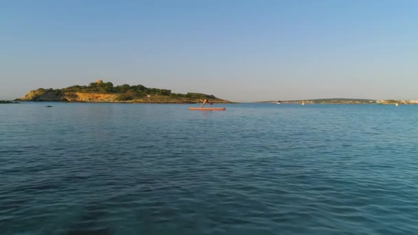 Tunnistamaton naissurffaaja uimapuvussa seisoo Adho Mukha Svanasanassa meressä melalaudalla ja harjoittelee joogaa auringonlaskun aikana - Materiaali, video