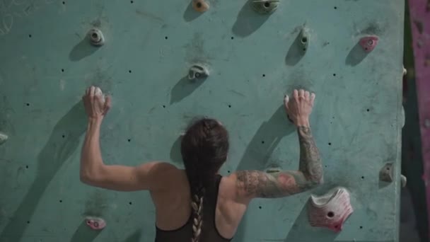 Vista posterior de atleta irreconocible tatuado poderosa mujer escalando en la pared colorida con repisas para escaladores en la habitación - Metraje, vídeo