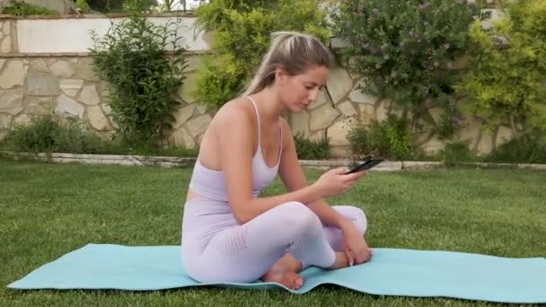 Spor giyim bölümünde sakin bir kadın yoga yaparken cep telefonunu kullanarak mindere çıplak ayakla oturuyor. - Video, Çekim