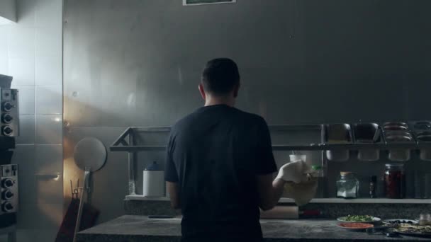 Rückansicht eines Mannes, der beim Zubereiten einer Pizza in der Nähe verschiedener Zutaten und Utensilien in der Pizzeria-Küche auf einem Marmortisch Stretching frischer Teig dreht und wirft - Filmmaterial, Video