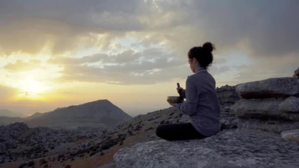 Vista laterale a basso angolo della tranquilla femmina seduta in posizione Padmasana con ciotola per cantare e meditare mentre si rilassa in montagna all'ora del tramonto - Filmati, video