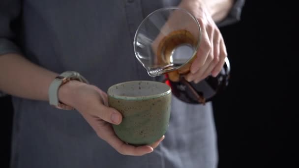 Crop anonyymi naaras kaatamalla tuoretta kuumaa keitettyä kahvia lasi syöttäjä keraaminen kuppi hidastettuna mustaa taustaa vasten - Materiaali, video