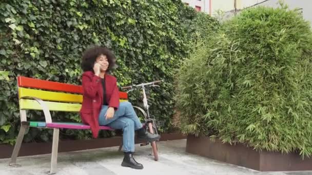 Содержание афроамериканка сидит на скамейке в городе и разговаривает на смартфоне, глядя в сторону - Кадры, видео