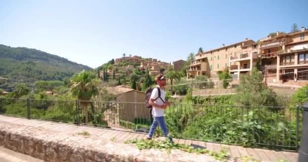 Teljes hossza elegáns szakállas férfi turista divatos ruhát és napszemüveg hátizsák séta a hídon az óvárosban található zöld dombok nyári napon - Felvétel, videó