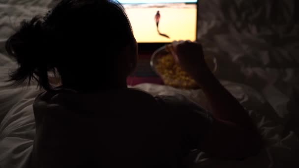 Z góry nierozpoznawalna pani w piżamie leżąca na łóżku z miską popcornu oglądając wieczorem film - Materiał filmowy, wideo