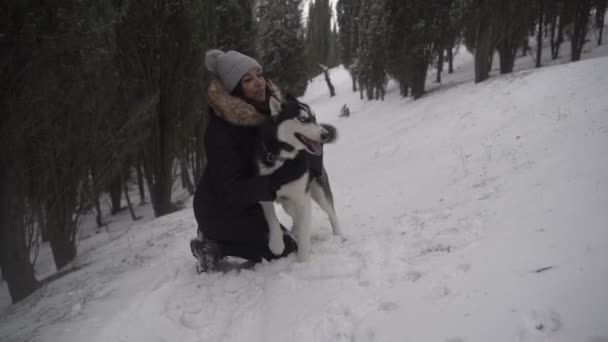 Улыбающаяся молодая женщина в верхней одежде обнимает и целует милую хаски-дог, приседая в снежных лесах возле зеленых елок в зимний день - Кадры, видео