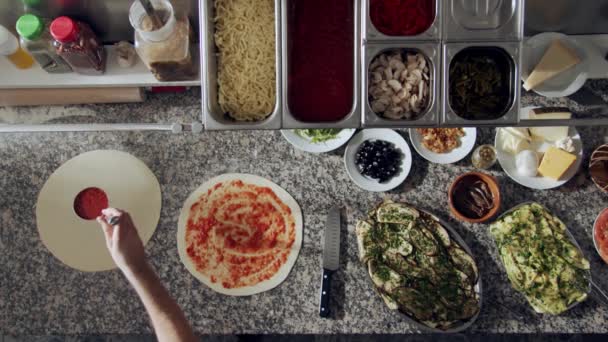 Bovenaanzicht van bijgesneden anonieme chef-kok gieten en verspreiden tomatensaus op deeg met lepel tijdens het koken pizza op marmeren tafel met verschillende ingrediënten in restaurant keuken - Video