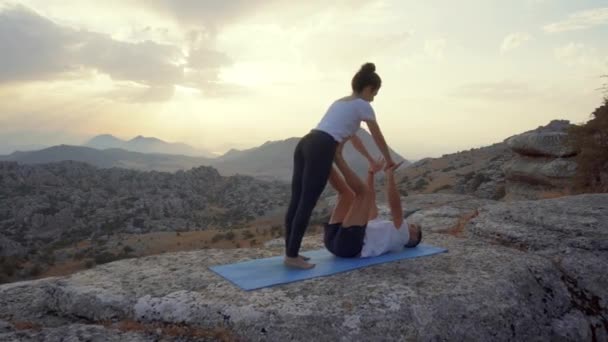 Vista lateral de cuerpo completo del hombre y la mujer enfocados practicando acro yoga y haciendo Front Plank asana en la cima de la montaña rocosa - Imágenes, Vídeo