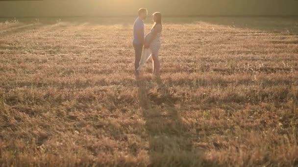 Вид збоку чоловіка м'яко тримає руки і цілує вагітну жінку, стоячи, дивлячись один на одного в сухому полі під час заходу сонця в сільській місцевості
 - Кадри, відео