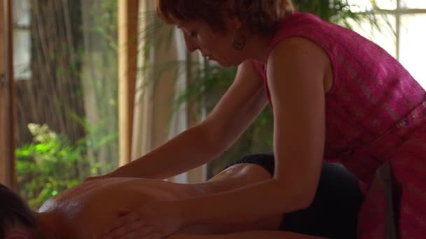 Vista lateral del maestro realizando masaje ayurvédico con aceite aromático en la espalda del cliente relajado en el salón moderno - Imágenes, Vídeo