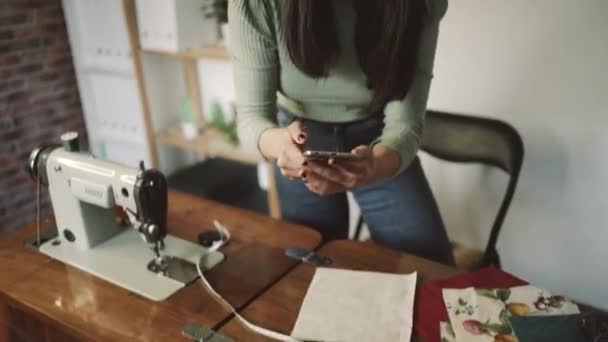 onherkenbare naaister praten met smartphone staande leunde naar voren tijdens het gebruik van rechte lijn aan tafel met papier en schaar in loft stijl studio - Video