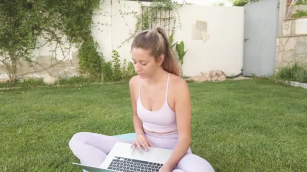 Mujer tranquila en ropa deportiva sentada descalza en la esterilla y eligiendo un tutorial en línea para practicar yoga - Imágenes, Vídeo