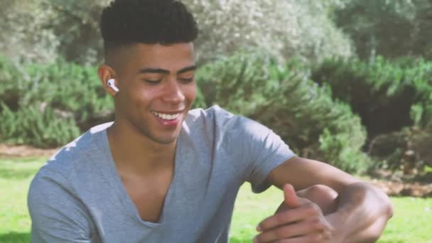 Jonge etnische man in sportkleding en draadloze oortelefoons lezen berichten op smartphone en grinniken tijdens het zitten op het gazon in de buurt van versnelling en rusten tijdens de training in het park - Video