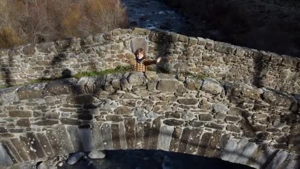 Aktywny zabawny dzieciak spacerujący po starożytnym moście łukowym pod małym górskim potokiem przez leśną dolinę w jasny dzień - Materiał filmowy, wideo