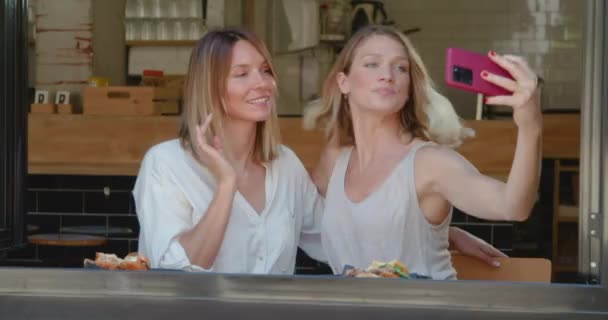 Expresszív barátnők grimaszolás közben selfie okostelefonnal kávézóban ül közel nyitott ablak - Felvétel, videó