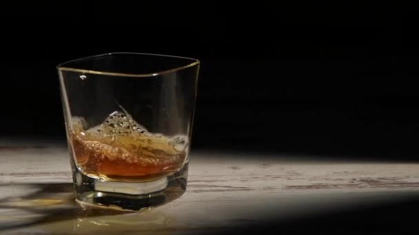Henkilö täyttää läpinäkyvän lasin bourbonilla puupöydällä säteillä mustaa taustaa vasten - Materiaali, video