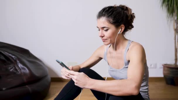 Adult content female athlete in earphones watching video tutorial on cellphone during training on mat in house - Footage, Video
