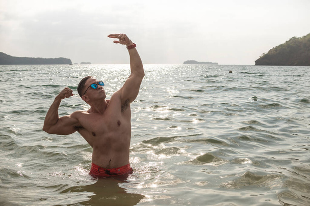 A fit and confident asian man does a classic aesthetic bodybuilding pose waist deep in the water. - Photo, Image