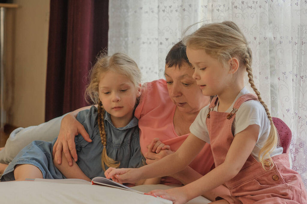 Η γιαγιά διαβάζει ένα βιβλίο σε δύο εγγονές στο σπίτι. - Φωτογραφία, εικόνα