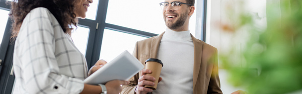 Hombre de negocios sonriente con café mirando a un colega afroamericano con tableta digital, pancarta  - Foto, Imagen