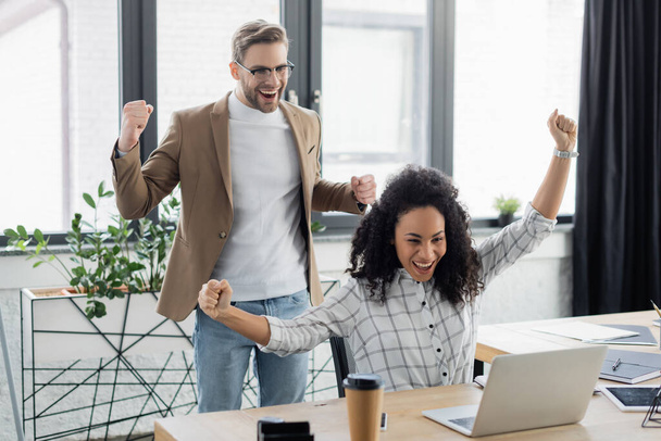 Empresários inter-raciais excitados mostrando sim gesto perto de laptop no escritório  - Foto, Imagem