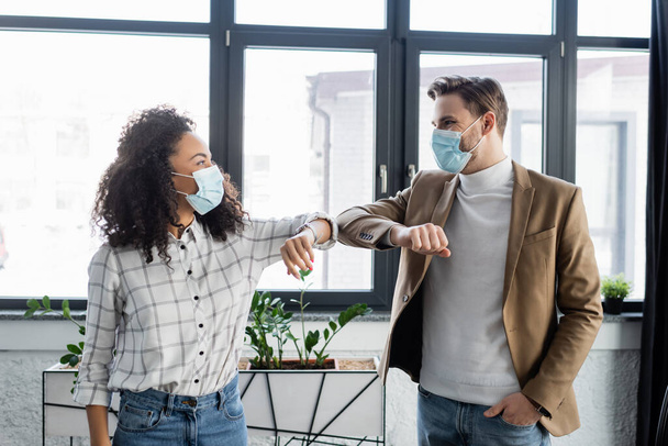 junge interrassische Geschäftskollegen in medizinischen Masken beim Ellbogenschlag im Büro - Foto, Bild