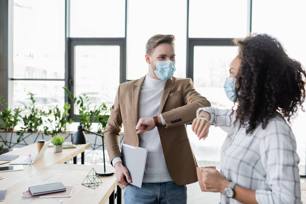 interracial business partners in medical masks doing elbow greeting in office - Photo, Image