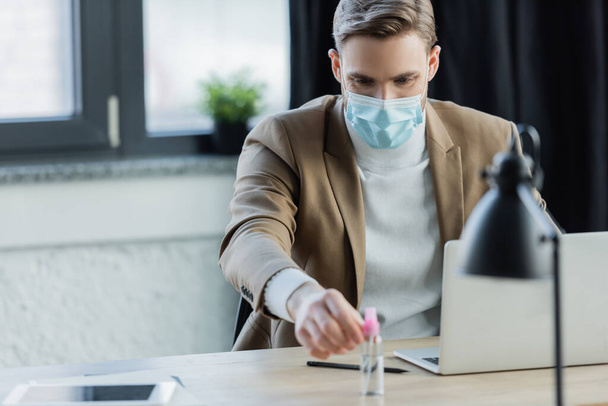 młody biznesmen w masce medycznej biorąc antyseptyczne siedząc w miejscu pracy - Zdjęcie, obraz