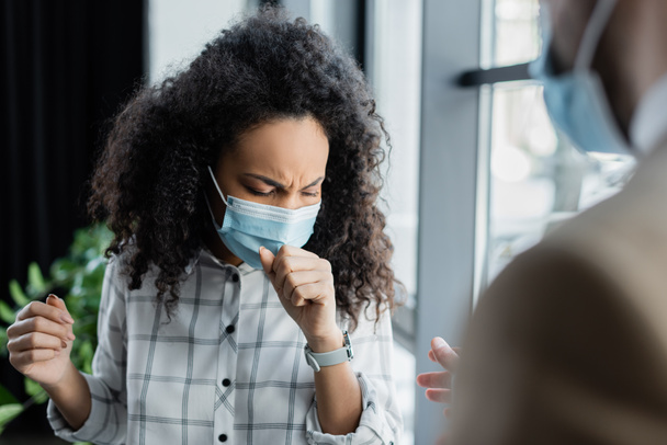 mujer de negocios afroamericana enferma con máscara médica tosiendo cerca de su compañero de trabajo en primer plano borroso - Foto, Imagen