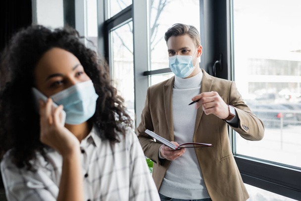 hombre de negocios en máscara médica con cuaderno y pluma cerca de colega afroamericano hablando en el teléfono inteligente, borrosa primer plano - Foto, imagen