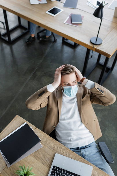visão de alto ângulo do empresário cansado em máscara médica que sofre de dor de cabeça no escritório - Foto, Imagem