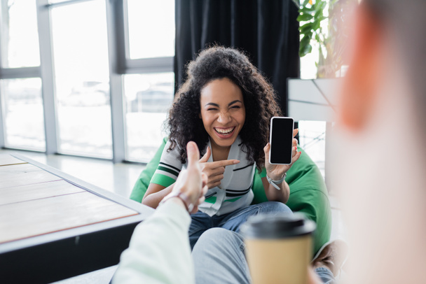 alegre afroamericana mujer apuntando al teléfono móvil con pantalla en blanco cerca de socio de negocios en primer plano borrosa - Foto, Imagen
