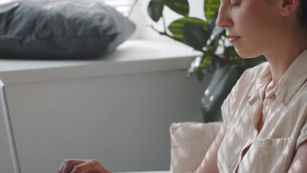 Inclinazione colpo di giovane donna seduta in poltrona a casa, tenendo il computer portatile in grembo e digitando - Filmati, video